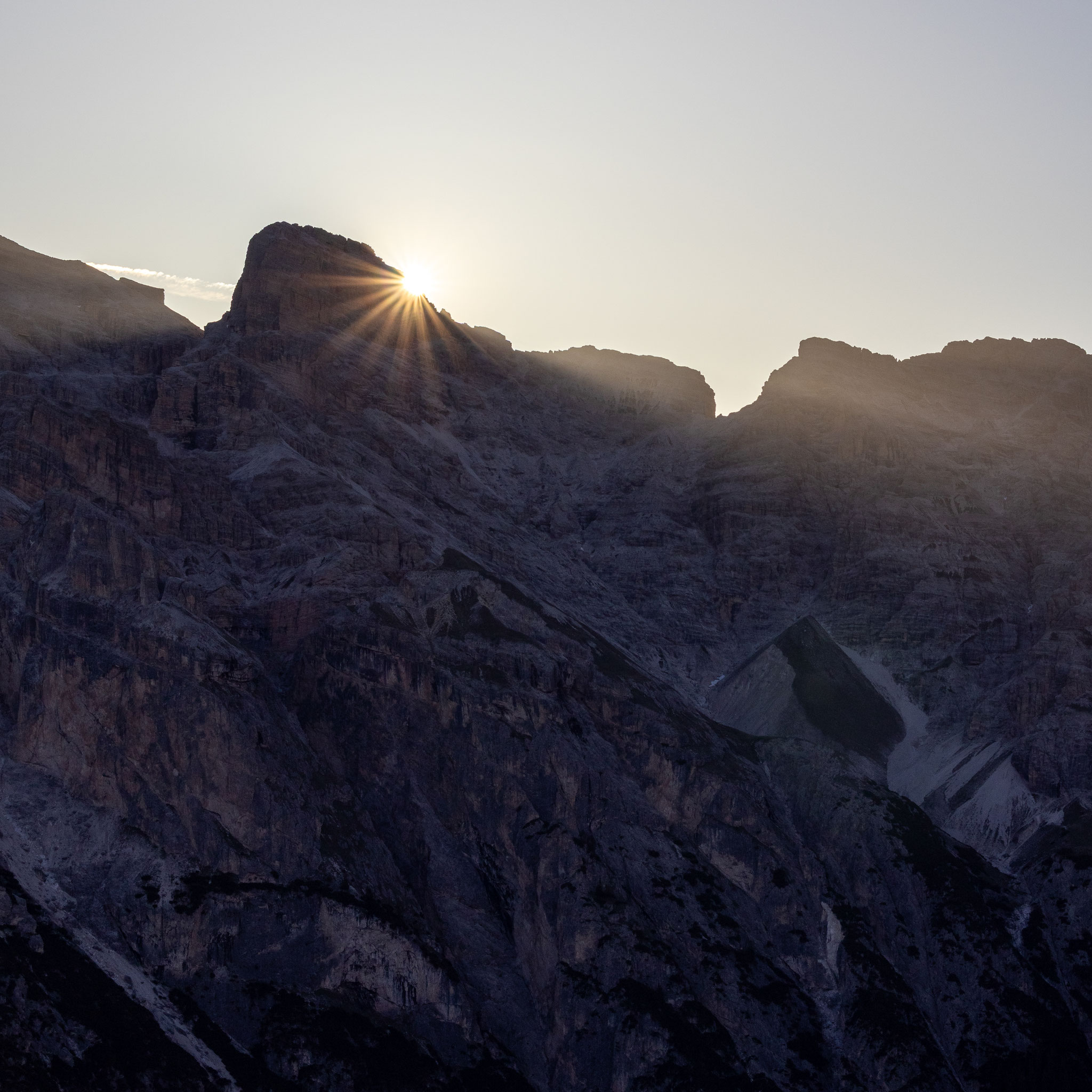 Die aufgehende Sonne bricht als heller Stern hinter einem Felsgrat der Dolomiten hervor und taucht die schroffen Bergwände in sanftes Morgenlicht. Die ruhige Szenerie vermittelt die besondere Stimmung eines Sonnenaufgangs in den Bergen.