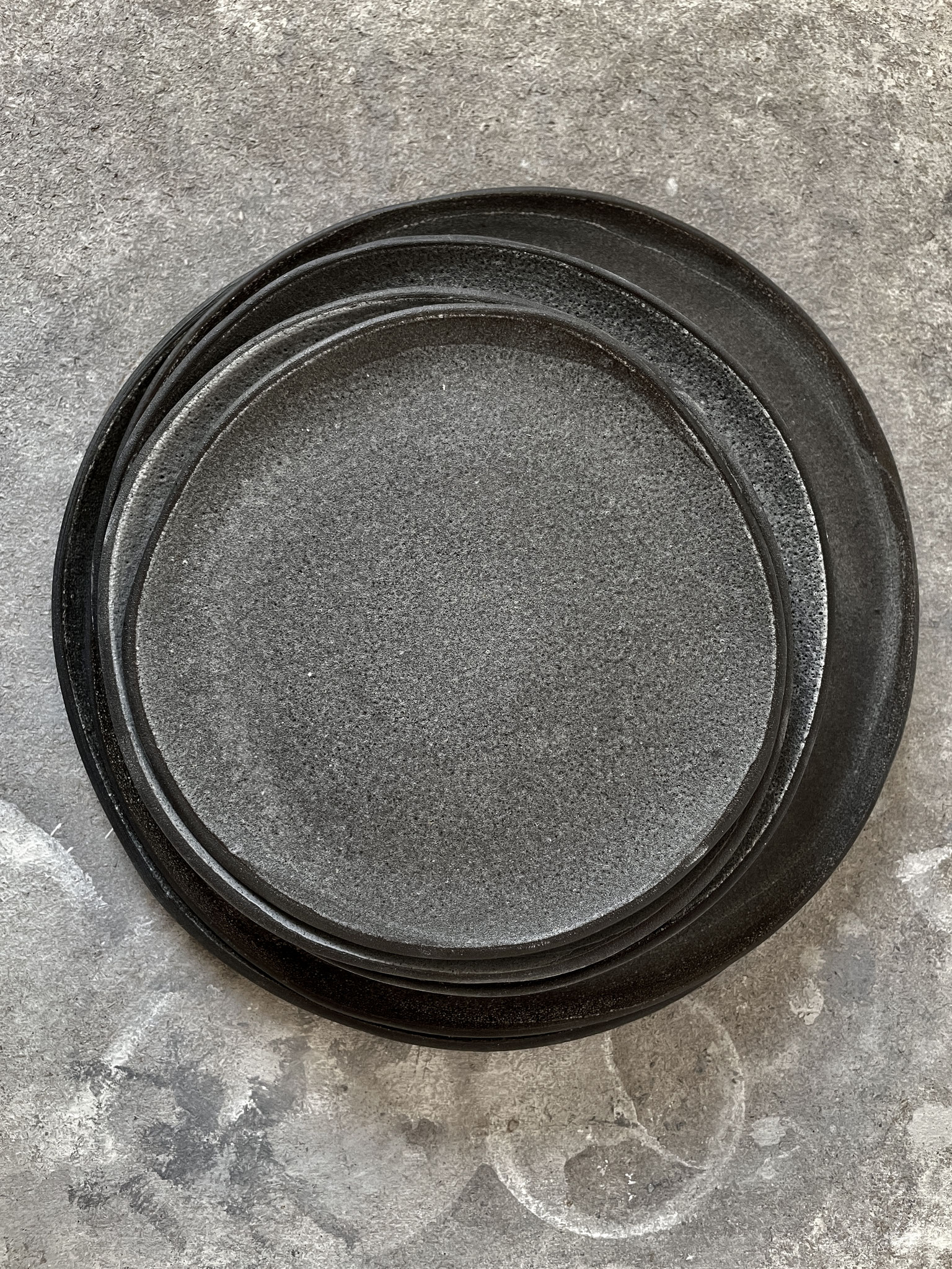 Ein Stapel Teller aus schwarzem Steinzeug-Ton aus der caldera serie. Schwarz, matt-milchiges design. 