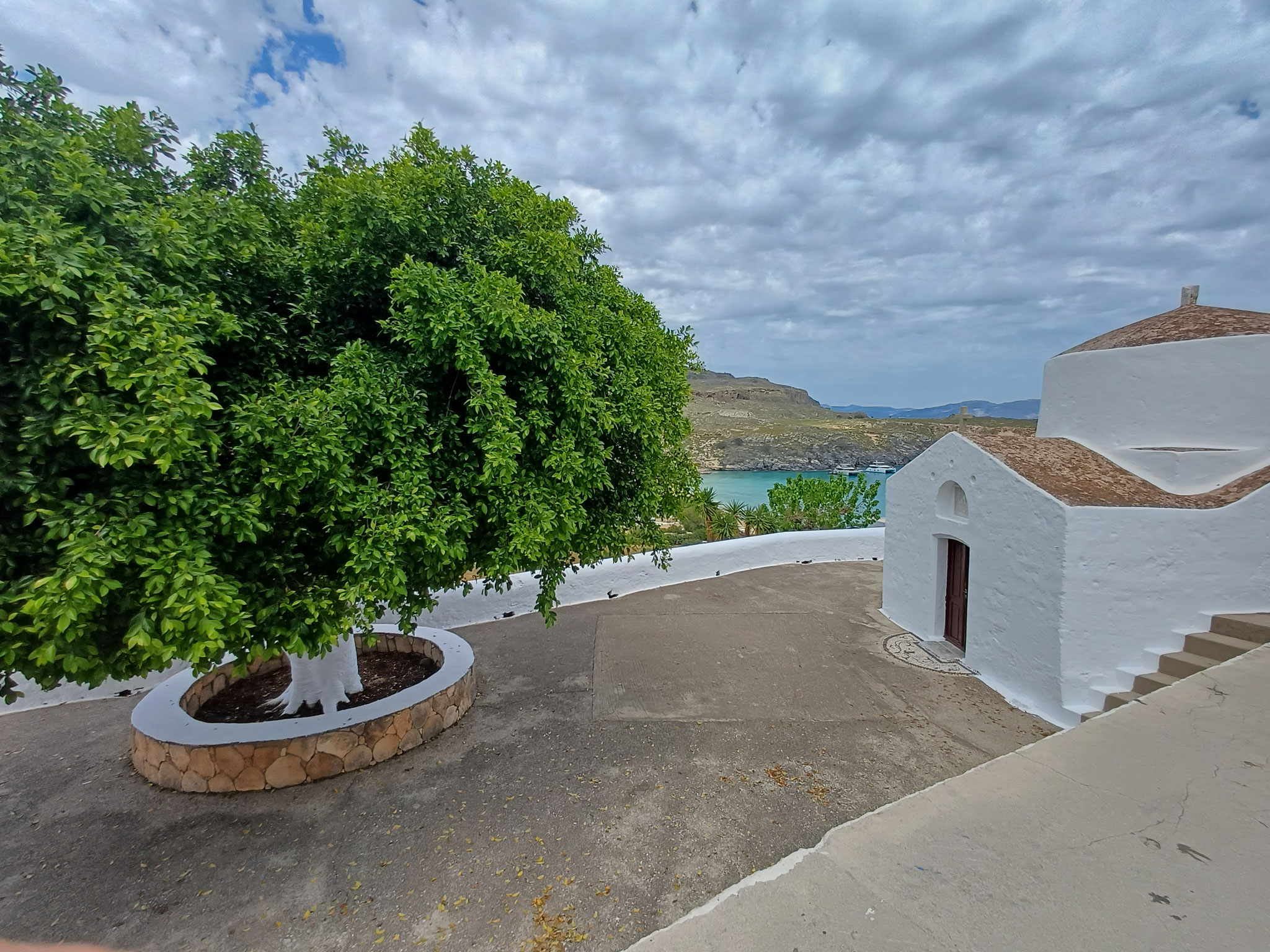 Ausblick auf einen Vorplatz. Auf der linken seite befindet sich ein großer Baum. Auf der rechten Seite ist eine weiß gestrichene Kirche zu erkennen.