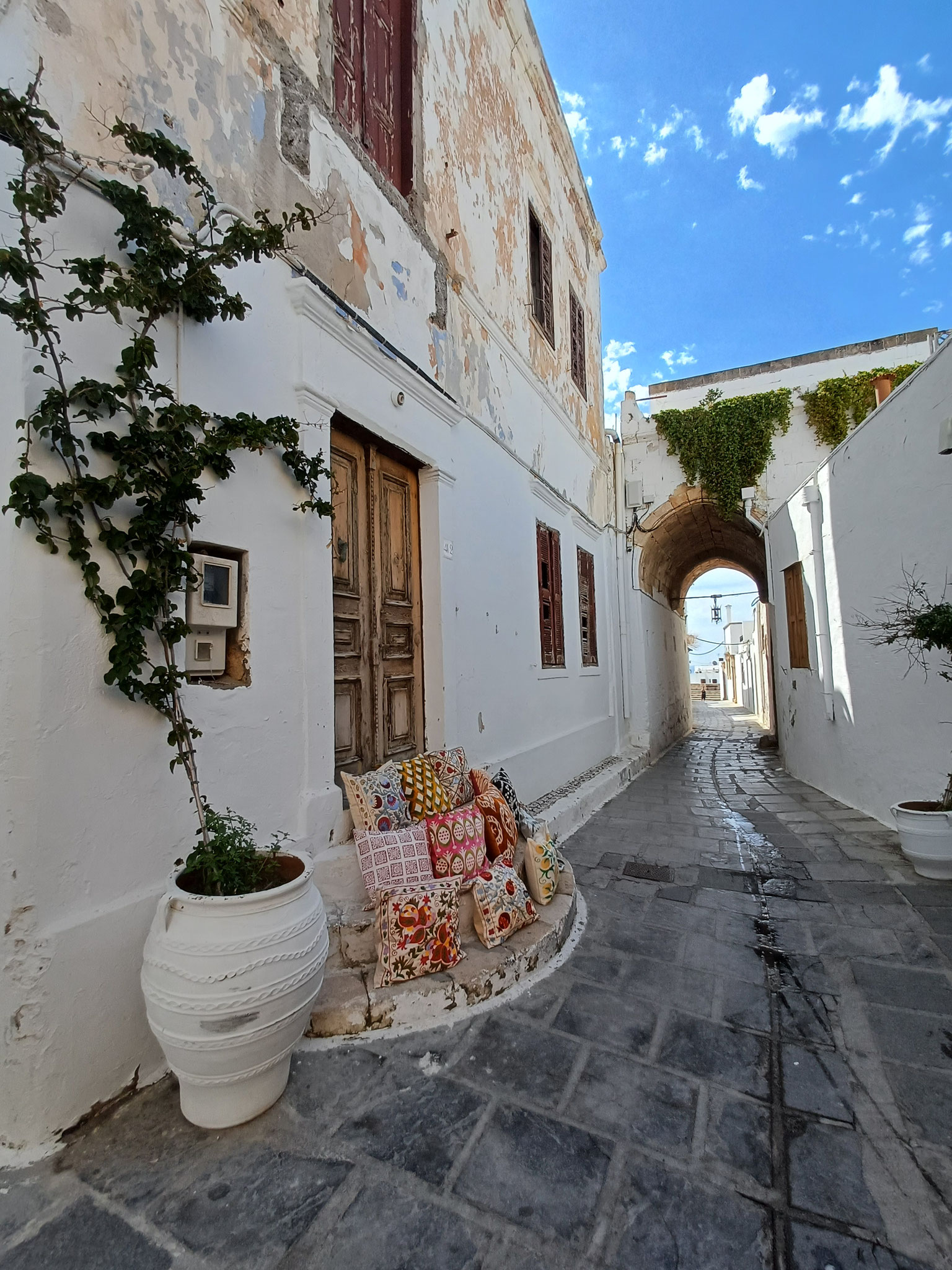 Schmale Gasse umgeben von weiß bemalten Häusern mit Rundbogen im Hintergrund. Vor der Eingangstür auf der linken Seite liegen verschiedenfarbige Kissen und ein Pflanze.