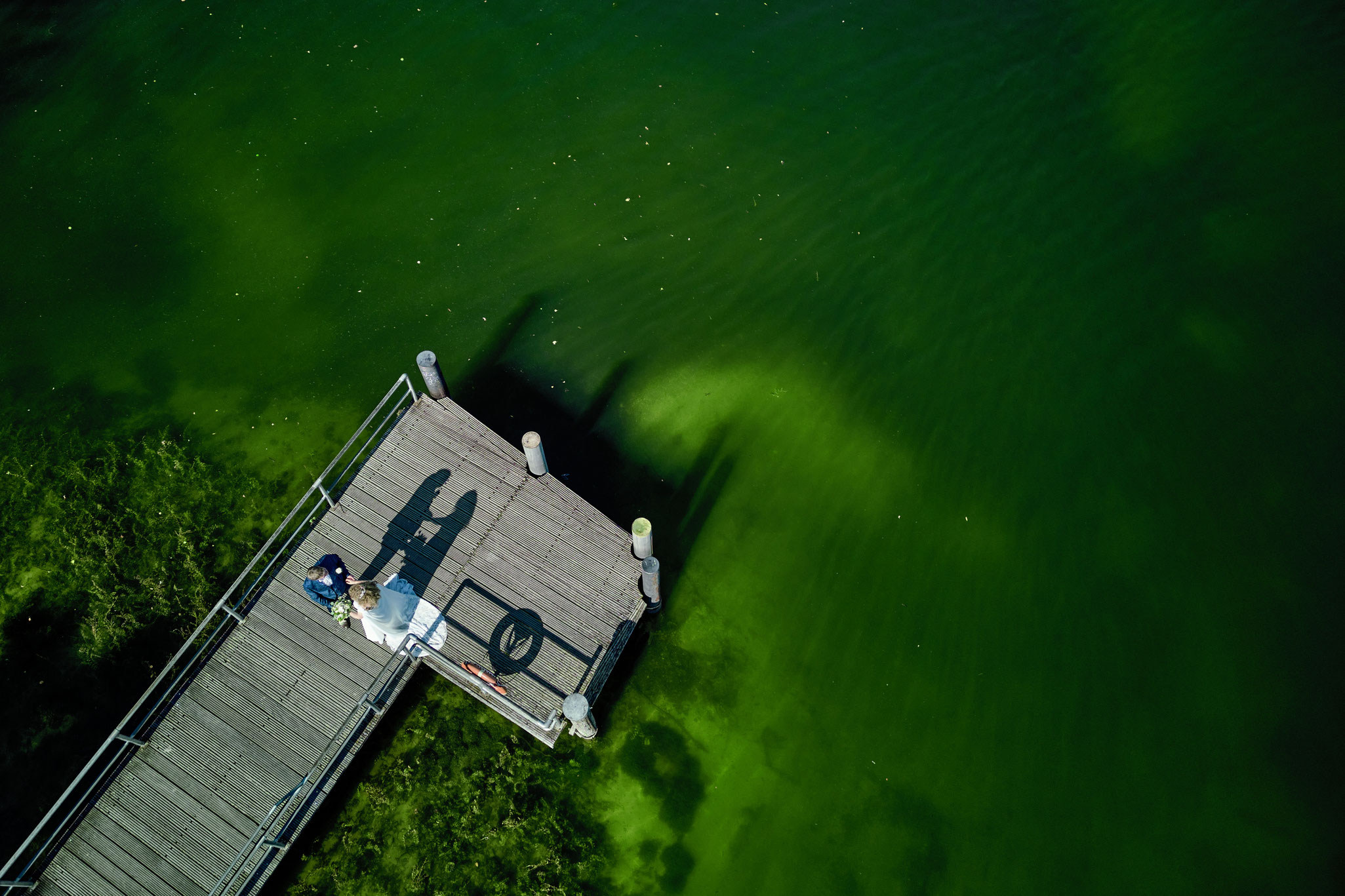 Drohnenaufnahme eines Brautpaares auf einem Steg auf dem Plöner See, das Wasser der Umgebung ist fast unwirklich grün
