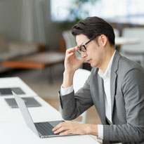 ノートパソコンの前で疲れを感じて眉間を押さえる中年男性会社員