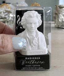 Eine Hand, von der man den Fingernagel des Daumens sehen kann, hält den Beethoven-Radierer in seiner durchsichtigen Verpackung. Der Beethoven-Radierer ist winzig, die Verpackung edel. Es ist in einem Wohnumfeld arrangiert.