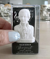 Eine Hand, von der man den Fingernagel des Daumens sehen kann, hält den Mozart-Radierer in seiner durchsichtigen Verpackung. Der Mozart-Radierer ist winzig, die Verpackung edel. Es ist in einem Wohnumfeld arrangiert.