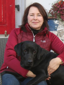 Sabine sitzt lächelnd auf einem Stuhl. Ein schwarzer Labrador ist mit Kopf und Pfoten auf ihrem Schoss.