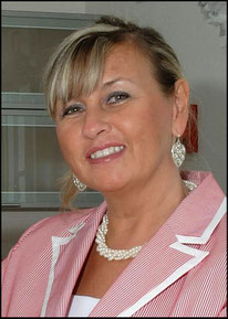 The portrait of the owner of the BACH 4 YOU Publishing House, Renate Bach. She smiles to the viewer. She has a red and white jacket on and wears a perl necklace