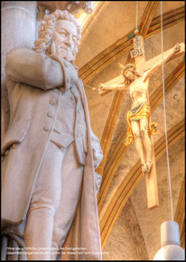 It is a photo of a Bach monument inside a church. In the background the is Jesus on the cross. The atmosphere is a warm yellow tone.