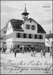 A historic town hall in the middle of a German village. In front of it there a persons. It's an old photography.