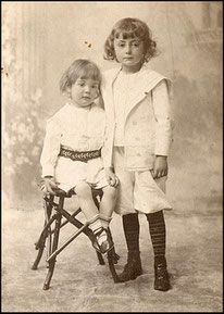 On a historic black and white yelowish photo there are two children. One is sitting on a chair. They are looking to the viewer and they are not smiling.