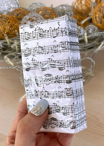 A hand with a glittering fingernail holds a packet of paper handkerchiefs. They are white and have black notes and staves on them. It is wrapped in foil. There are home decorations in the background.