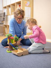Logopädin und ein kleines Mädchen führen eine spielerische Sprachübung mit Therapiematerial auf dem Boden während einer logopädischen Behandlung in einer Praxis in Villach durch.