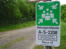 Das Foto zeigt neben einer Forststraße das offizielle Hinweisschild mit der Bezeichnung des Rettungstreffpunkts.