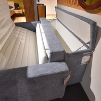 Graues Schlafsofa mit hochgeklappter Rückenlehne, die Stauraum für Bettzeug freigibt.