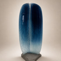 Grande sculpture en verre bleu de Prusse, translucide aux lignes verticales, installée sur une base minérale.