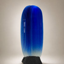 Sculpture contemporaine en verre bleu electrique, bleu klein,  translucide aux lignes verticales, posée sur un socle minéral sombre.