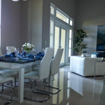 design project by Mia Home Trends featuring a staged modern living and dining area with white dining table, white dining chairs, white sofa and chair, glass tables and blue accents
