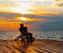 Auf einem Bootssteg am Meer sitzt ein Angler. Am Abendhimmel ist die untergehende Sonne teils von dunkleren Wolken verdeckt.