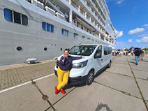 White van parked on dock in front of cruise ship in Riga port with woman in purple blouse, yellow trousers and red shoes