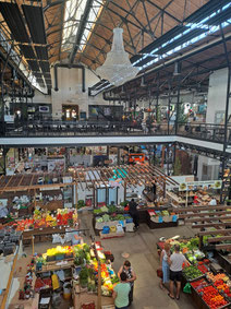 Inside the pavillion of Āgenskalns Market in Rig with colourful produce stalls on the ground floor and a large chandelier hanging from the ceiling 