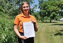 Eine duale Studentin in orangenem T-Shirt zeigt eine DIN-A4-große weiße Unterlage zur Kamera und lächelt dabei. Im Hintergrund sind Wiese, Obstbäume und große Bäume