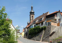 Man sieht eine kleine Straßenschlucht in einem Boxberg. Auf der rechten Straßenseite ist eine Wand, die zum Teil begrünt ist, danach folgen Wohnhäuser und ein Kirchturm.