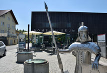 Am Gebäude im Hintergrund steht in großen Buchstaben RÖMERMUSEUM. Links sieht man ein Haus, rechts die Außenfläche eines Cafes. Dominant steht im Vordergrund ein Kunstwerk aus Blech: ein Römer in Rüstung, mit Helm, Schild und Pilum.