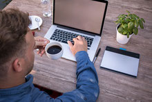 Ein Mann sitzt vor einem Laptop, eine Hand ist auf der Tastatur, die andere Hand führt eine Tasse schwarzen Kaffee zum Mund. Neben dem Laptop liegen ein Block und ein Stift auf dem Tisch, daneben steht ein kleiner Topf mit einer Grünpflanze