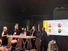 Tina Barroso moderiert die Diskussionsrunde zur Biodiversität im Solarpark im Rahmen des BNEW Sommerfests mit vier Referentinnen und Referenten. Alle stehen an einem Podiumstisch. Im Hintergrund eine Grafik auf der Leinwand.