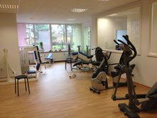Fitnessraum mit verschiedenen Trainingsgeräten wie Crosstrainer, Rudergerät, Hantelbank und Kletterwand, helle Fenster und Holzfußboden