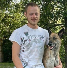 Chris steht im Freien vor Bäumen und trägt ein weißes T-Shirt mit dem Schriftzug 'Bullet For My Valentine'. Er hält seinen Hund Tio, der braun-weiß gefleckt ist und aufmerksam nach rechts schaut.