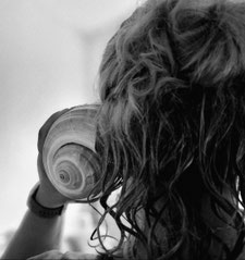 Schwarz-weiß Bild von einer Person von hinten. Man sieht einen Hinterkopf mit längeren gelockten Haaren, die Person hält sich eine große Muschel ans Ohr. 
