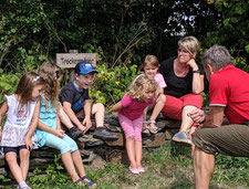 fünf Kindergartenkinder sitzen auf einer Natursteinmauer und hören einem Herrn in rotem Poloshirt zu. Neben den Kindern sitze eine mittelalte Frau - eine Kita-Erzieherin, die den Kindern zusieht.  