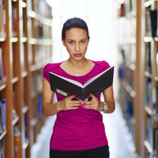 Eine Frau steht zwischen den Regalen einer Bibliothek und hält ein aufgeschlagenes Buch in den Händen