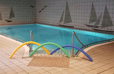 Innen-Schwimmbecken mit blauen Wasser, umgeben von weißen Fliesen, bunte Schwimmnudeln in Gelb, Grün und Blau liegen am Beckenrand, Wand mit Segelboot-Motiven