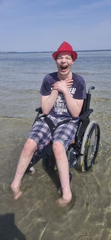 Auf dem Bild ist ein fröhlicher Moment bei einem Tagesausflug ans Meer festgehalten. Man sieht einen jungen Mann im Rollstuhl, der ausgelassen lacht und seine Hände vor Freude an die Brust gelegt hat. Seine Füße stehen im Wasser.