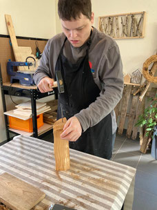 Auf dem Bild ist ein junger Mann in der Tagesförderstätte bei der Holzbearbeitung zu sehen. Konzentriert arbeitet er mit Hammer und Meißel an einem Stück Holz, das er auf dem Tisch fixiert hat. Er trägt eine schwarze Arbeitsschürze.