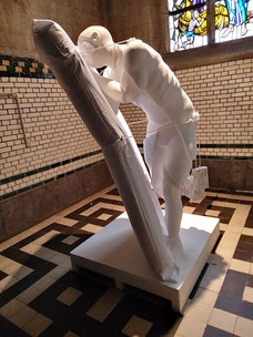 ‘The Image Generator’, Tentoonstellingszicht met witte slapende figuur die voorover leunt tegen een rechtopstaande matras; sculptuur op sokkel in betegelde ruimte met glasraam, Kunsthal Extra City, Antwerpen, 2023..