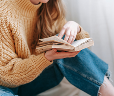 Nahaufnahme einer Frau in Jeans und einem dicken beigefarbenen Strickpullover, die ein aufgeschlagenes Taschenbuch in den Händen hält. Der Bildausschnitt konzentriert sich ganz auf das Buch, ihren Oberkörper, ihre Arme und Beine.