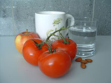 Mandeln, Tomaten und ein Apfel liegen vor einer Teetasse und einem gefüllten Wasserglas.