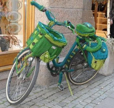 Ein Fahrrad mit Fahrradkorb und Fahrradtasche am Gepäcksträger ist vollständig mit einem gestrickten Mantel umhüllt.