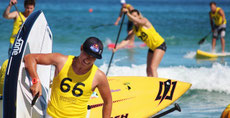 Mehrere Paddler in Shirts mit Startnummern paddeln in der Brandung und laufen mit Board und Paddel in den Händen über den Strand.