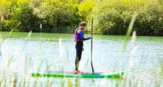 Auf einem See, umgeben von Natur steht ein Paddler in meditativer Pose auf seinem Board.