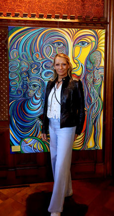 Zeitgenössische Kunst von Alexandra Mohr: im Vordergrund Alexandra Mohr, hinter ihr, ihr buntes großformat Acrylbild mit drei Gesichtern die sich in Schlaufen und Schlingen auflöst und in körperähnlichen Wesen manifestiert. Ausgestellt in Schloss Sinzig