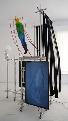Thermiekbelofte — installatie met een metalen frame op wielen waarin zwarte linten door een ventilator in beweging worden gebracht, een zwevende transparante figuur in een geometrisch skelet en een scherm waarop de 3D-animatie Mee met de wind wordt getoon