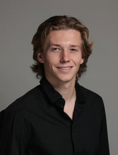 Maurice in einen schwarzen Hemd, vor einem grauen Hintergrund. Die Haare von Maurice sind blond/braun und ca. 10 cm. lang. 