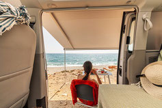 Eine Frau sitzt am Meer vor ihrem Wohnmobil gut geschätzt mit einem Camper Sorglos-Paket der TravelSecure