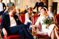 Un couple de mariés échange un regard complice et un tendre geste lors de leur cérémonie civile, entouré de leurs proches.