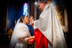 Une mariée reçoit la communion lors de son mariage religieux, dans une église éclairée par la lumière des vitraux.