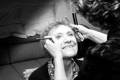 Une femme âgée sourit pendant qu’une maquilleuse applique son maquillage avec délicatesse, capturant un moment d’émotion et de préparation.