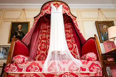 Une robe de mariée blanche suspendue à un lit à baldaquin rouge dans une pièce élégante, capturant l'élégance et l'anticipation du grand jour.